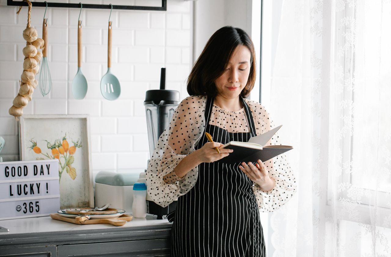 Asian woman in kitchen apron writing in a notebook. Perfect for culinary and lifestyle use.
